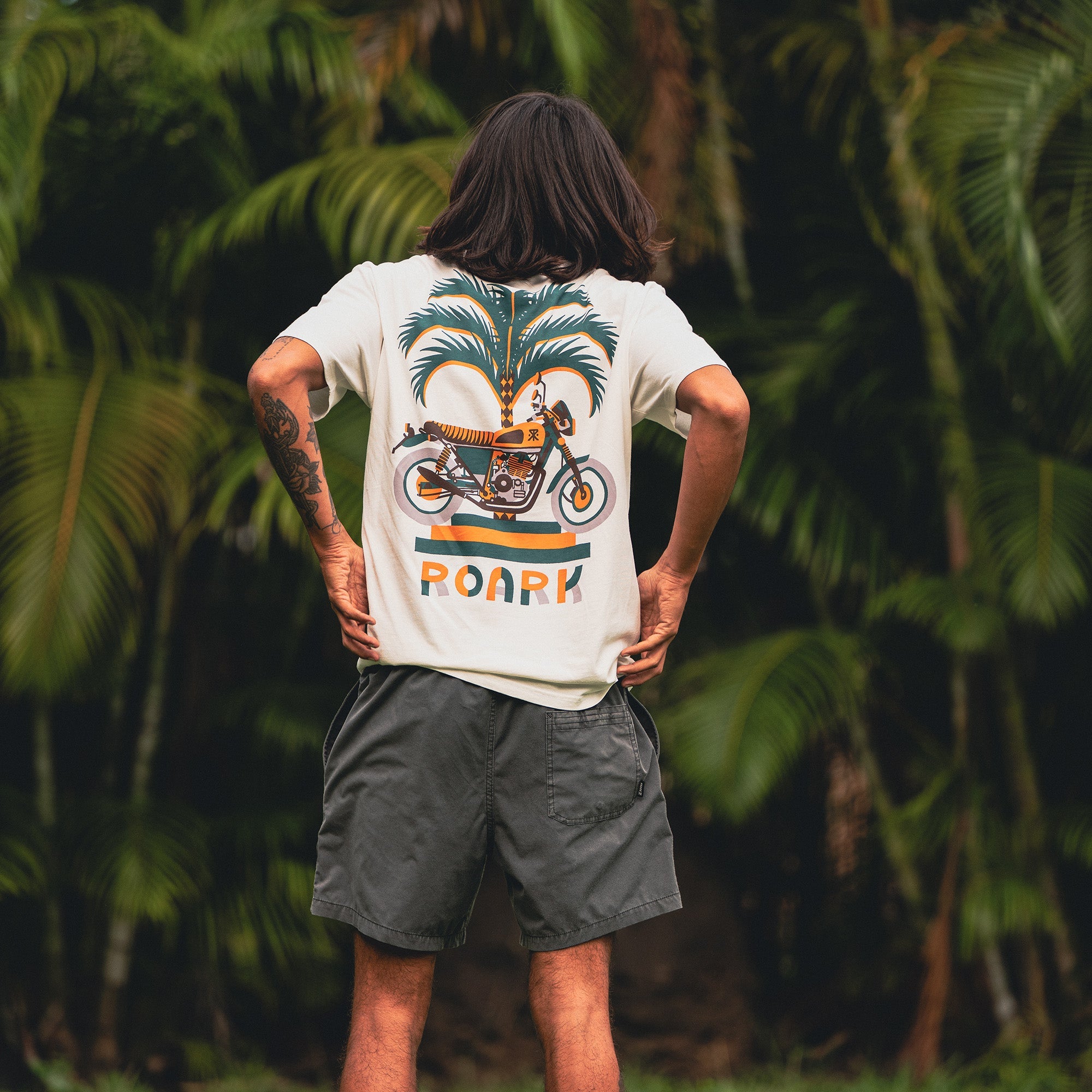 Man dressed in Roark Moto Palm Premium Tee and boardshorts, showcasing casual adventure-ready style with focus on sleeve and waist details.