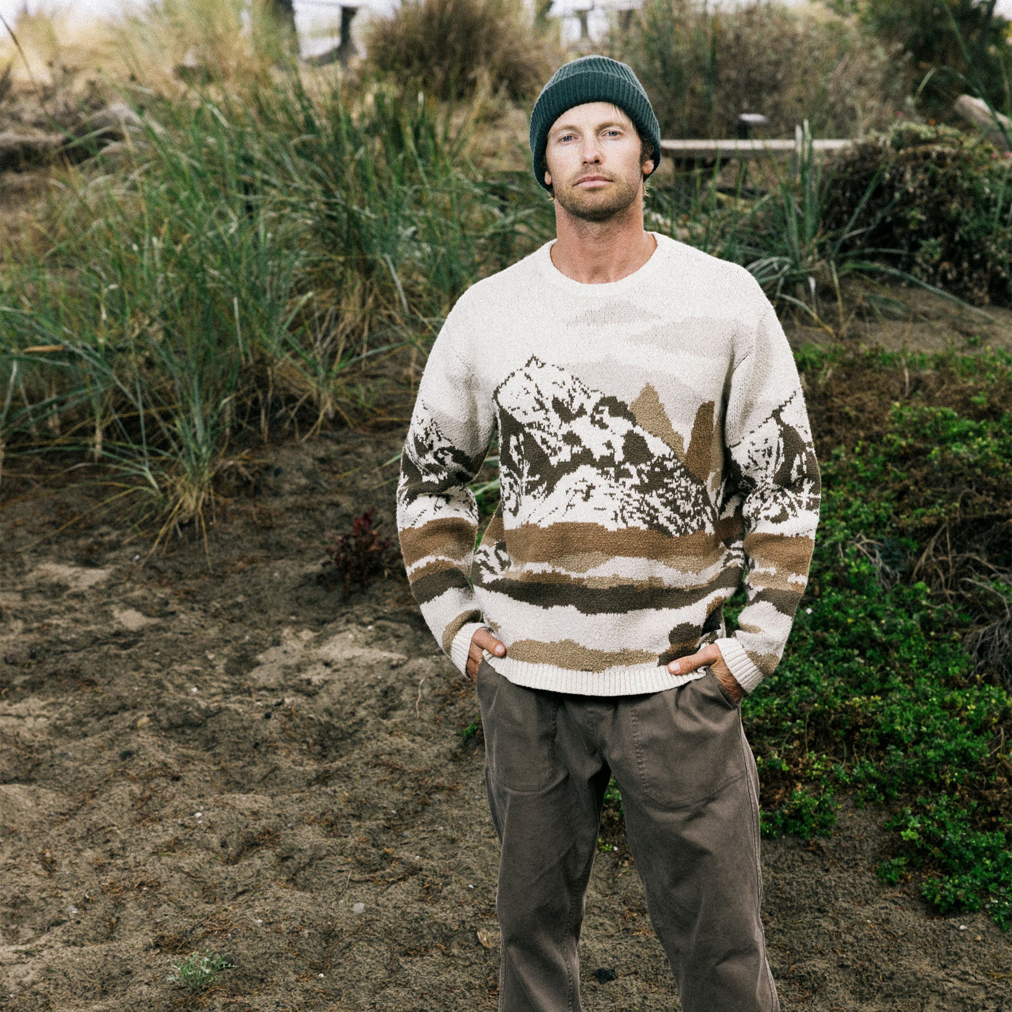 Man wearing a patterned sweater and beanie standing in a natural setting with plants and dirt ground.